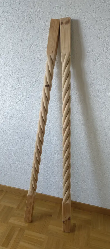 Man sieht zwei lange Säulen aus Fichtenholz, die an einer Wand lehnen. Sie sind oben und unten eckig. In der Mitte sind sie geschnitzt wie ein gedrehtes Seil.