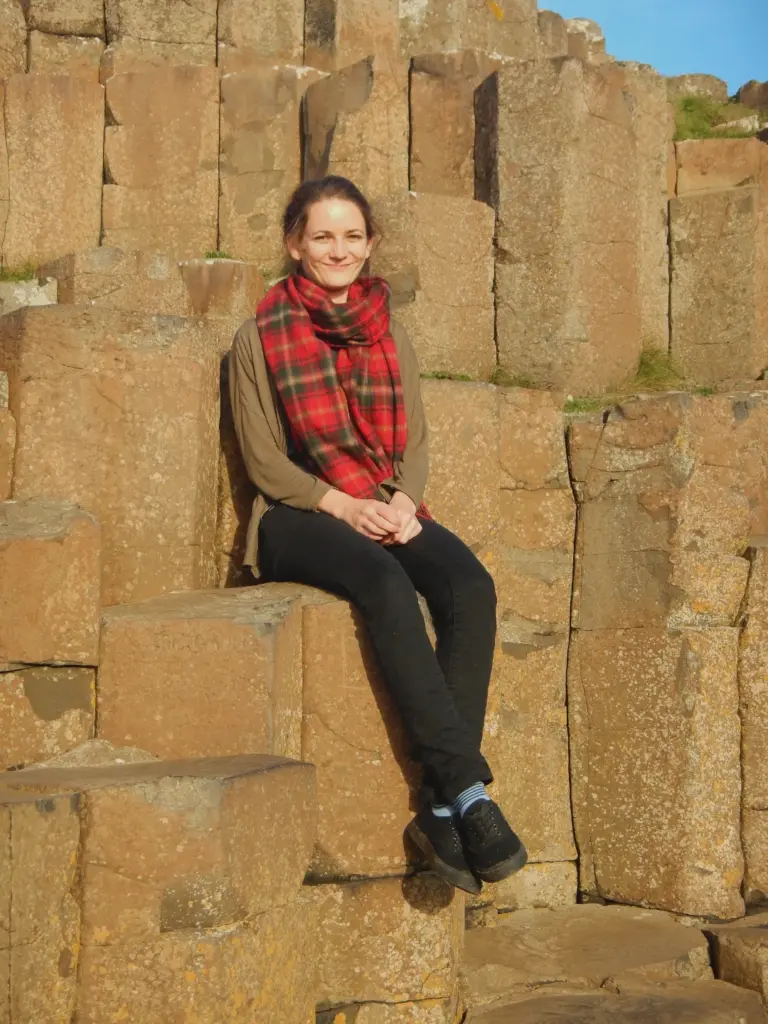 Blog03_Lisi Lisi sitzt auf einer der Basaltsäulen am Giant's Causeway in Irland. Sie trägt einen langen, roten Schal mit Tartanmuster.