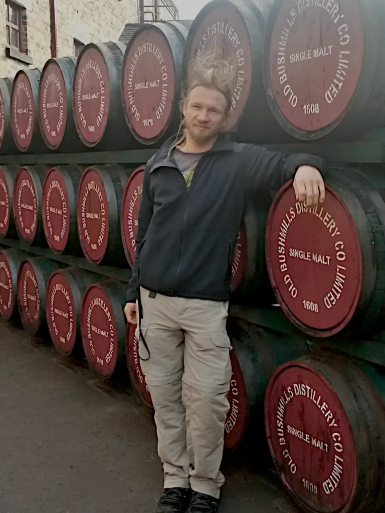 Blog03_Thomas Thomas steht vor einer Wand aus Whiskeyfässern auf dem Gelände der Bushmills Destillery. Seine Haare stehen nach oben, weil es sehr windig ist.