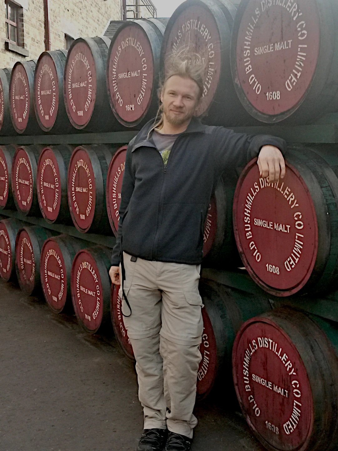 Blog03_Thomas Thomas steht vor einer Wand aus Whiskeyfässern auf dem Gelände der Bushmills Destillery. Seine Haare stehen nach oben, weil es sehr windig ist.